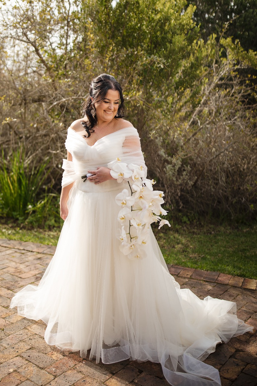 kim Ruched Tulle A-Line Gown with Off-the-Shoulder Elegance  The Kim gown is a show-stopping statement of soft structure and modern romance. Designed with layers of shimmer tulle and delicate ruching, this off-the-shoulder beauty features a flattering drop waist and dreamy movement. The wide horsehair hemline adds volume and definition, making every step unforgettable.  ✨ Design Highlights: ✔ Ruched Tulle Bodice – Sculpted to flatter with soft, elegant folds and a supportive fit. ✔ Off-the-Shoulder Straps – Romantic draped straps that highlight the collarbone and shoulders. ✔ Drop Waist Silhouette – Elongates the torso for a modern, flattering shape before flowing into a soft A-line. ✔ Shimmer Tulle Layers – A subtle sparkle throughout the skirt adds a radiant glow. ✔ Ruched Tulle Bolero – Optional matching ruched tulle bolero for added coverage or drama. ✔ Wide Horsehair Hemline – Creates structured movement and a crisp, couture finish to the skirt. ✔ Zip & Button Back Closure – Smooth fit with classic bridal button detailing.  💫 Customization Options: ✅ Custom fit to your exact measurements ✅ Option to remove bolero or adjust strap style ✅ Available in ivory, soft white, or blush shimmer tulle ✅ Train length and hemline width can be customized  💎 Kim is made for the bride who wants soft glamour, modern sophistication, and a dash of drama on her special day. 💎  📞 Book your fitting or custom consultation today! 📧 info@bestersbridal.co.za | 📱 084 268 1354  Ruched Tulle • Drop Waist • Off-Shoulder Straps • Shimmer Layers • Horsehair Hem • A-Line Silhouette