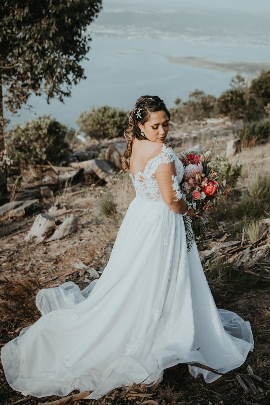 GILLIAN A captivating white A-line bridal gown made from layers of soft tulle and adorned with intricate lace. The sheer lace bodice features delicate long sleeves, offering a touch of romance, while the low back bodice adds a hint of allure. The full-flare A-line skirt flows gracefully, embellished with lace appliqués that cascade down to the wide horsehair hemline, adding structure and volume for a striking silhouette. This gown blends timeless elegance with modern detailing, perfect for a bride seeking both grace and charm on her special day.