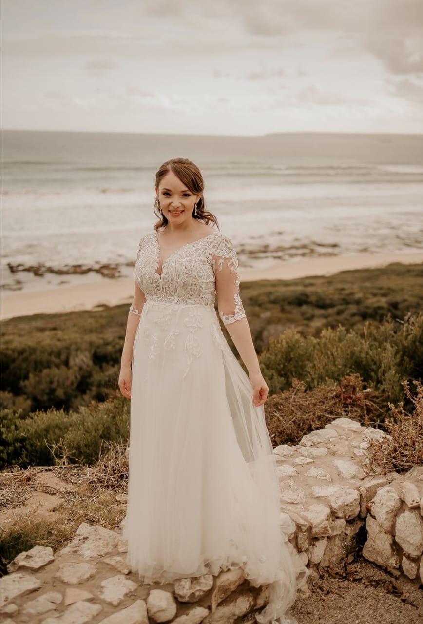 MELANTHA A romantic tulle and lace A-line wedding gown that epitomizes timeless beauty. The sheer lace bodice showcases intricate lace detailing, offering a delicate yet elegant touch. Complementing the design are 3/4 lace sleeves that provide a refined and sophisticated appeal. The flowing A-line tulle skirt is adorned with exquisite lace appliqués, adding texture and charm to the silhouette. A subtle short train completes the look, along with a classic button closure for a graceful finish. Perfect for the bride who desires a blend of elegance and comfort.