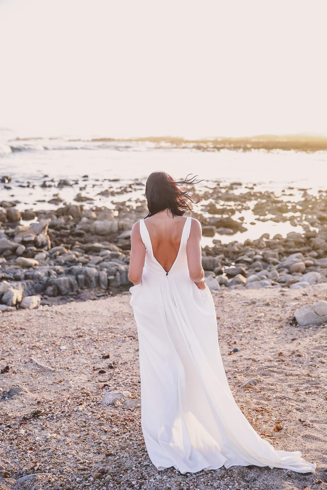 ANNEKE This stunning off-white A-line dress, crafted from smooth crepe, exudes effortless elegance, making it a perfect choice for a beach wedding. The flattering V-neckline adds a hint of allure, beautifully balancing the simplicity of the design, while the low V-back bodice creates a modern and romantic silhouette. Functional side pockets provide a stylish yet practical touch, ideal for a carefree, beachy vibe. The full-flare skirt flows gracefully, allowing for easy movement and a dreamy, windswept look as you walk along the shore. This gown perfectly captures a laid-back yet chic bridal style.