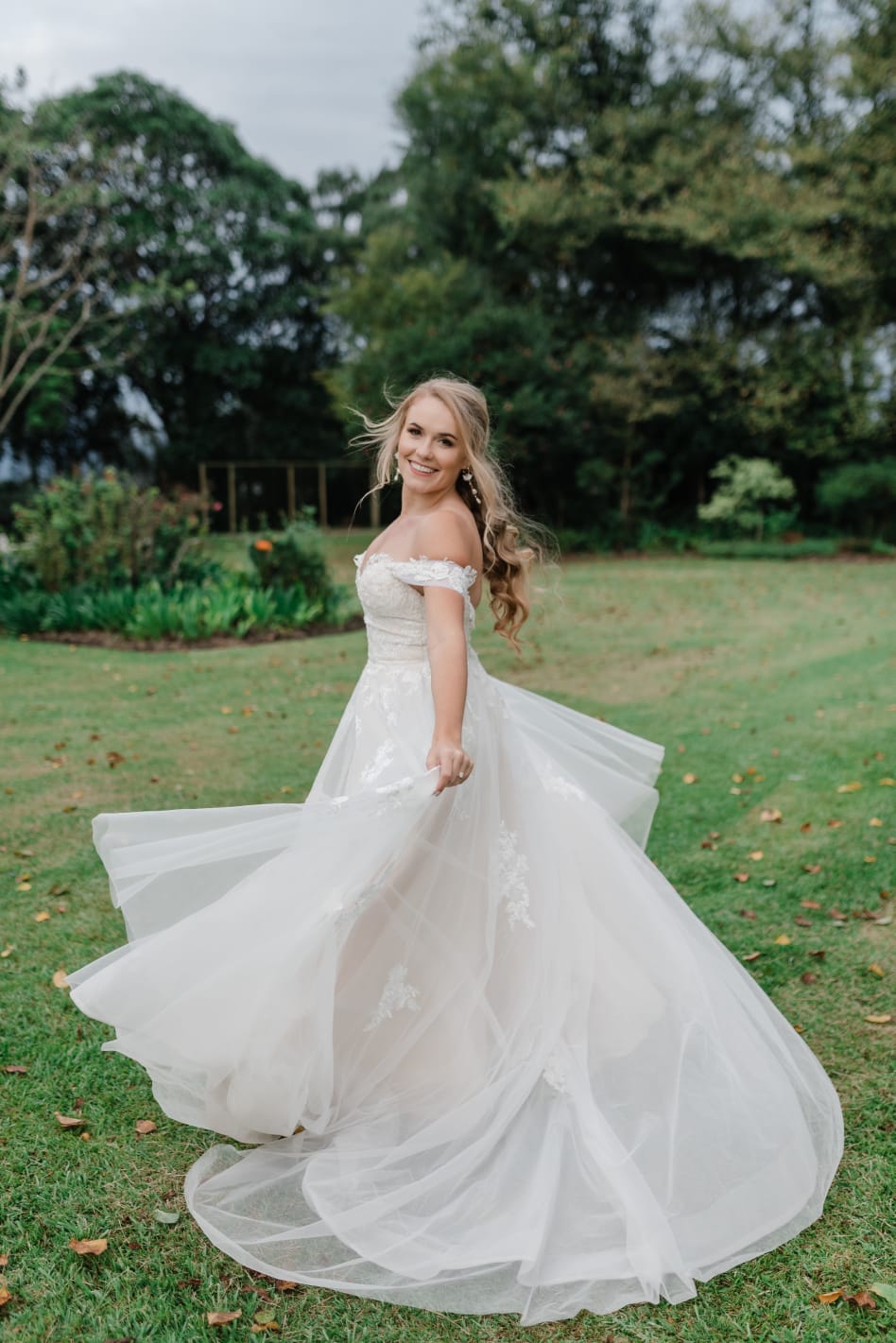 HAILEY A dreamy blush wedding gown, designed with shimmering tulle and delicate lace in a soft blush hue. The strapless sweetheart neckline beautifully frames the décolletage, while the bodice is adorned with intricate floral lace appliqués that add texture and romance. The A-line tulle skirt cascades gracefully, embellished with floral lace details that create a stunning, whimsical effect. Detachable draped tulle sleeves offer a versatile touch, allowing the bride to switch between a classic strapless look and an effortlessly romantic style. This gown captures the perfect balance of elegance and modern bridal charm.