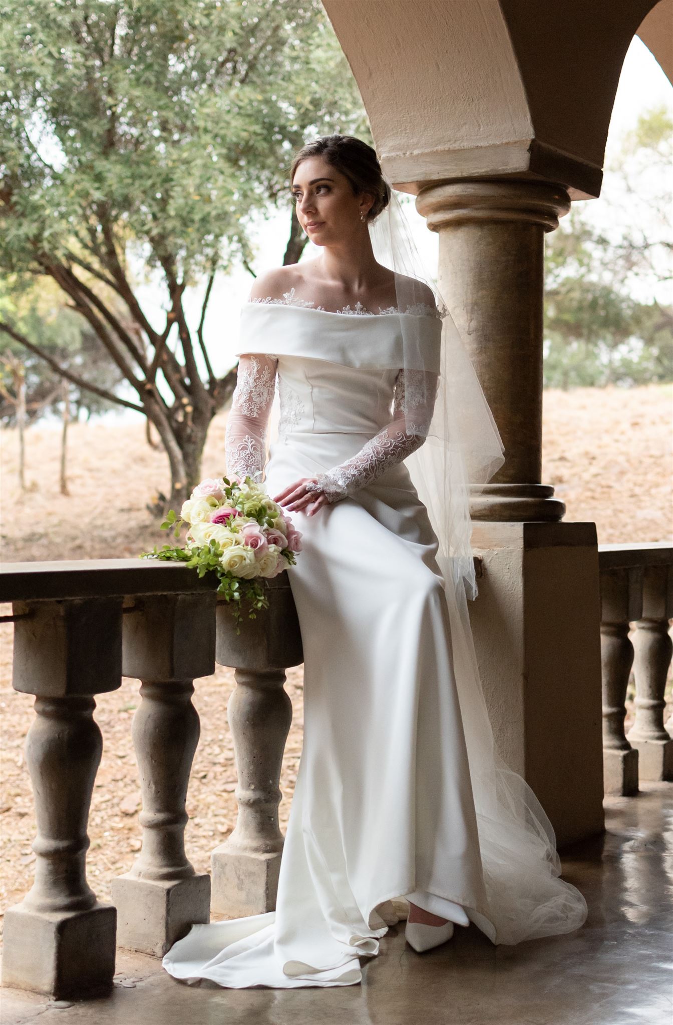 Dress Maxine : A sophisticated off-the-shoulder sheath bridal gown crafted from smooth crepe fabric, adorned with intricate corded lace appliqués. The gown features sheer lace long sleeves that add a touch of elegance, with lace detailing gracefully framing the neckline. The back boasts a delicate button closure, leading to a subtle short train, creating a refined and timeless look. The dress is available for custom orders, tailored to your exact measurements in the colour of your choice. You can personalize it further by selecting either a long or short train, with short or long sleeves, and with or without a slit.