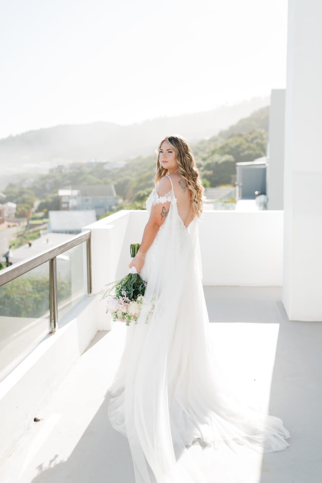 CHeynaid An ethereal off-white bridal gown, crafted from layers of delicate tulle and lace. The gown features a captivating deep plunge sheer lace bodice, adorned with intricate lace appliqués that subtly accentuate the bride’s figure. The A-line tulle skirt flows effortlessly, adding a touch of whimsy and romance, while a daring thigh-high slit offers an unexpected modern twist. Completing the look are detachable draped tulle sleeves that cascade gracefully, giving the bride the option to add a soft, romantic flair or embrace a sleek, strapless silhouette for a more versatile style.