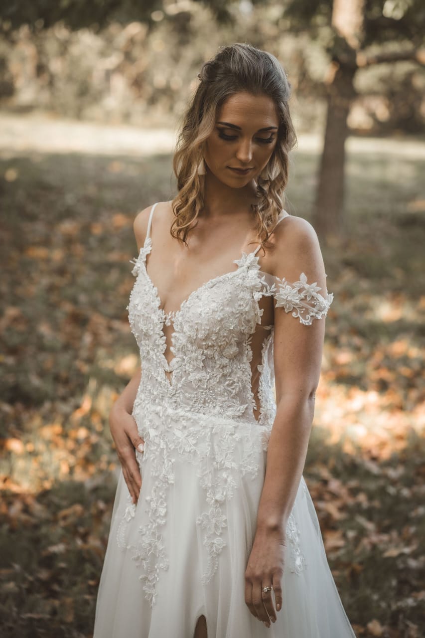 MARELIZE This enchanting bridal gown features a deep plunging V-shaped neckline crafted from luxurious off-white Chantilly lace and adorned with delicate embroidered lace details. The bodice is supported by thin, elegant straps and complemented by gracefully draped off-the-shoulder lace straps for a romantic touch. The gown flows into a stunning A-line tulle skirt, designed with a daring high slit that adds a contemporary edge to its timeless elegance. The back showcases a dramatic low V-shaped design, ensuring breathtaking views from every angle.